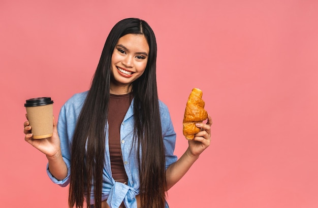 Retrato de uma jovem e bela mulher asiática com fome comendo croissant Retrato isolado de mulher com fast food e café sobre fundo rosa Conceito de café da manhã de dieta