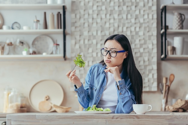 Retrato de uma jovem e bela mulher asiática sentada na cozinha à mesa segurando um verde fresco