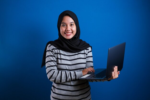 Retrato de uma jovem e bela mulher muçulmana asiática sorrindo enquanto segura um laptop, uma estudante bem-sucedida contra um fundo azul