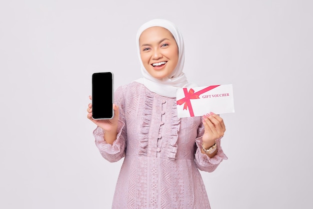 Retrato de uma jovem e bonita e alegre mulher muçulmana asiática usando hijab e vestido roxo segurando o vale-presente e mostrando o celular de tela em branco isolado no fundo branco do estúdio