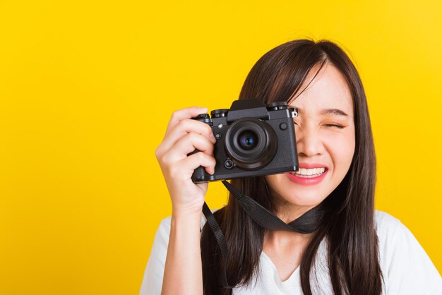 Retrato de uma jovem e bonita fotógrafa asiática sorridente tira uma foto e olha o visor em uma câmera fotográfica sem espelho digital retrô pronta para fotografar, tiro em estúdio isolado em fundo amarelo