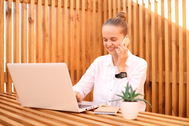 Retrato de uma jovem e bonita freelancer sorridente trabalha remotamente no laptop no café mulher falando no telefone inteligente no trabalho enquanto faz trabalho remoto no refeitório gosta de seu trabalho