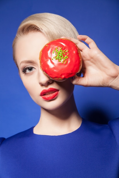 Retrato de uma jovem e bonita modelo com lábios vermelhos espreitando de um donut brilhante sobre fundo azul