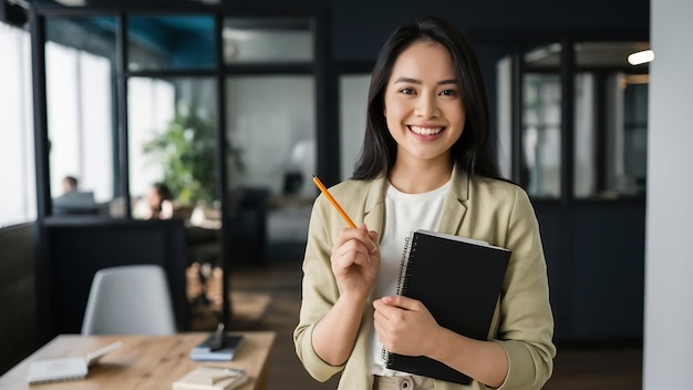 Foto retrato de uma jovem e bonita mulher asain segurando um caderno e um lápis enquanto está de pé em um escritório moderno