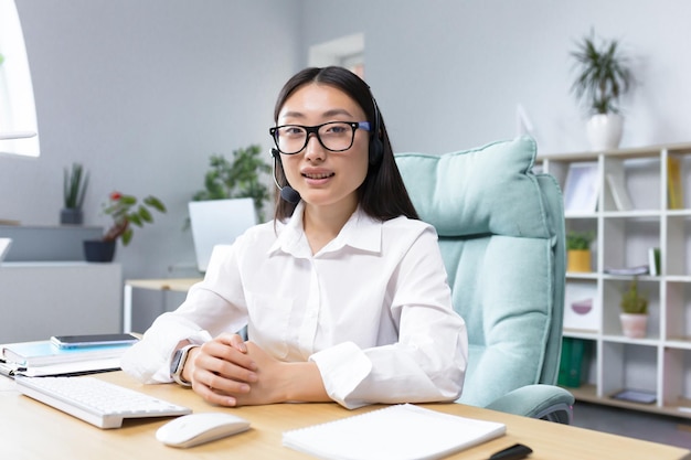 Retrato de uma jovem e linda mulher asiática em óculos e fones de ouvido com um microfone sentado no