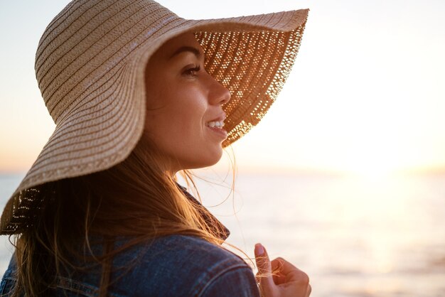 Retrato de uma jovem elegante e feliz em uma jaqueta jeans branca e um chapéu de palha na costa do oceano a.