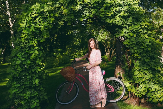 Retrato de uma jovem elegante em um vestido floral rosa longo, pare de andar sob o arco de carvalho em uma bicicleta com uma cesta para compras, comida ou flores ao ar livre, tempo de recreação feminino bonito na primavera ou no parque de verão