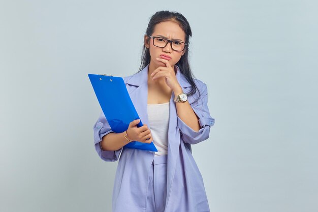 Retrato de uma jovem empresária asiática séria segurando o queixo, contemplando algo enquanto segura a pasta de documentos isolada em fundo roxo