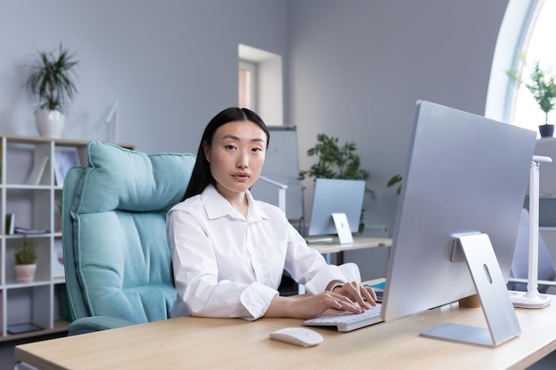 Retrato de uma jovem empresária bem-sucedida, uma trabalhadora em um escritório moderno trabalhando em um computador, uma mulher asiática olhando para a câmera