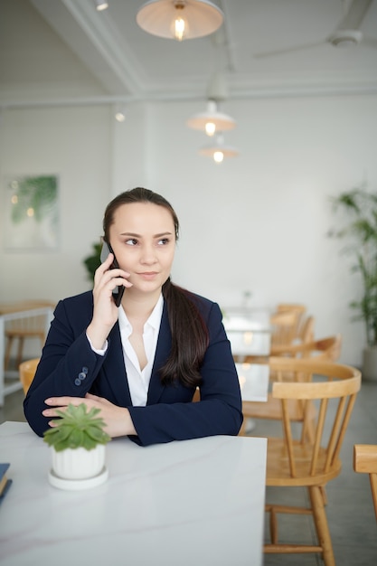 Retrato de uma jovem empresária positiva sentada à mesa do café e falando no telefone com um colega de trabalho