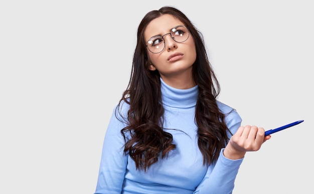 Foto retrato de uma jovem empresária séria vestindo blusa azul de manga comprida e óculos redondos transparentes trabalhador de escritório feminino europeu pensando e olhando para um lado na parede branca do estúdio