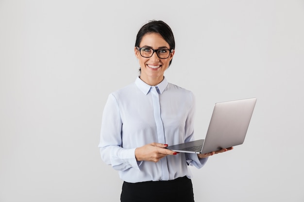 Retrato de uma jovem empresária usando óculos, segurando um laptop prateado no escritório, isolado sobre uma parede branca