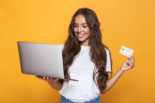 Retrato de uma jovem encantada com longos cabelos castanhos em pé sobre uma parede amarela, segurando um laptop, mostrando um cartão de crédito de plástico