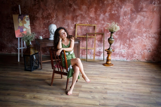 Retrato de uma jovem encantadora com cabelo encaracolado menina de pele clara em um vestido verde senta-se em uma cadeira e com um sorriso no rosto Mulher encaracolada em roupas leves