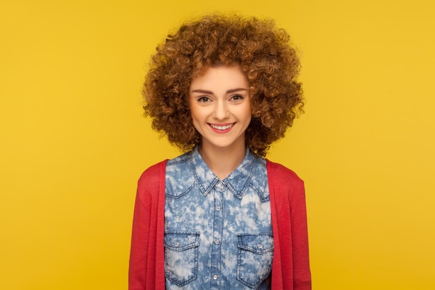 Retrato de uma jovem encantadora feliz com cabelos cacheados fofos em camisa jeans estilo casual, olhando para a câmera com um sorriso, regozijando-se com notícias positivas. tiro de estúdio interno isolado em fundo amarelo