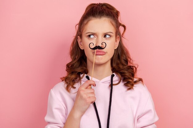 Retrato de uma jovem engraçada com capuz segurando o bigode de festa de papel preto na vara fazendo beicinho e olhando para o lado se divertindo nas férias tiro de estúdio interior isolado no fundo rosa