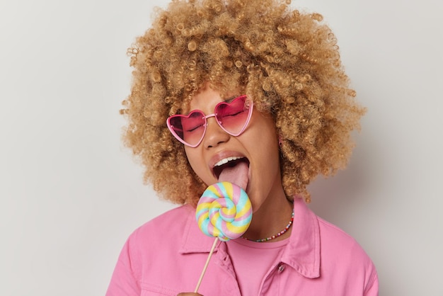 Foto retrato de uma jovem engraçada de cabelos encaracolados lambe doces multicoloridos de caramelo na vara usa óculos de sol cor-de-rosa da moda e jaqueta tem guloso isolado sobre fundo branco louco por doces