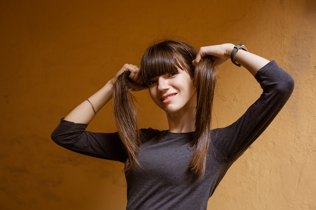 Retrato de uma jovem engraçada, segurando o cabelo nas mãos, olhando para a câmera contra uma parede laranja.