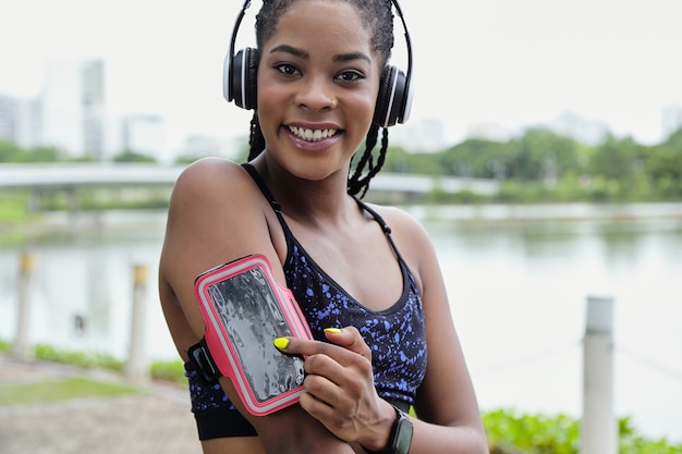Retrato de uma jovem esportista negra sorridente em fones de ouvido, apontando para a caixa do smartphone em seu braço