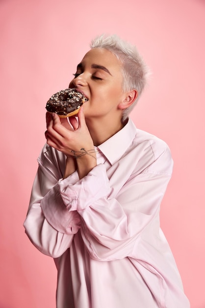 Retrato de uma jovem estilosa com cabelo curto posando comendo rosquinha isolada sobre fundo rosa Sobremesa