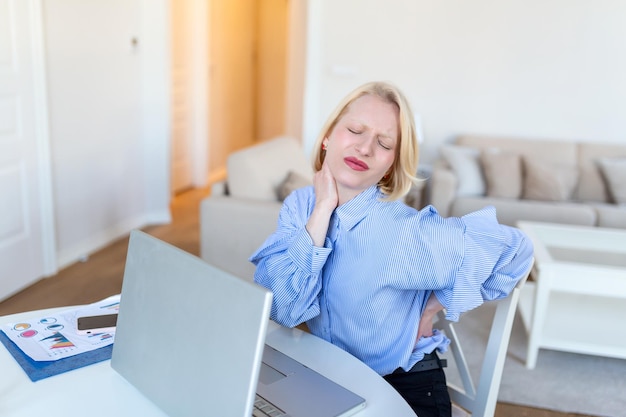 Retrato de uma jovem estressada sentada na mesa de escritório em casa na frente do laptop tocando as costas doloridas com expressão de dor sofrendo de dor nas costas depois de trabalhar no laptop