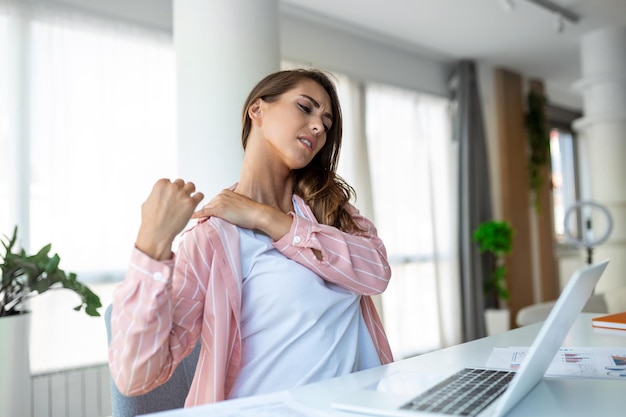 Retrato de uma jovem estressada sentada na mesa de escritório em casa na frente do laptop tocando o ombro dolorido com expressão de dor sofrendo de dor no ombro depois de trabalhar no laptop