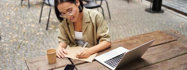 Retrato de uma jovem estudando on-line sentada com um laptop escrevendo, fazendo anotações e olhando para