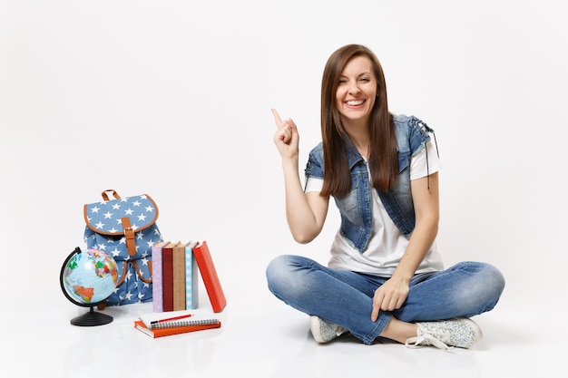 Retrato de uma jovem estudante alegre em roupas jeans, apontando o dedo indicador para cima, sentada perto do globo, mochila, livros escolares isolados