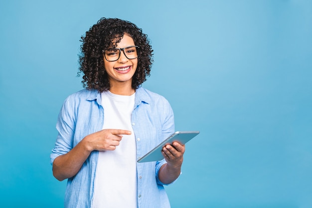 Foto retrato de uma jovem estudante americana com cabelo encaracolado africano segurando um tablet digital e sorrindo em pé sobre um fundo azul isolado