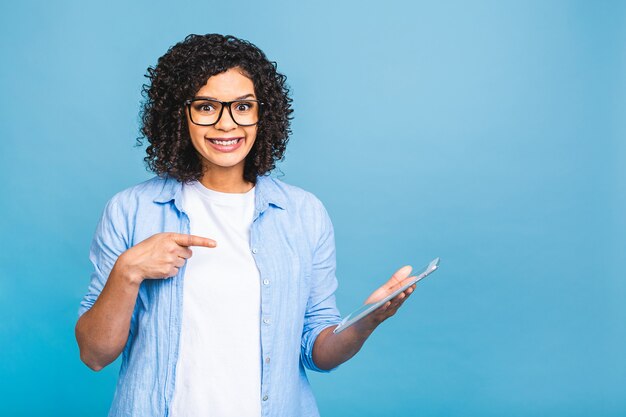 Foto retrato de uma jovem estudante americana com cabelo encaracolado africano segurando um tablet digital e sorrindo em pé sobre um fundo azul isolado