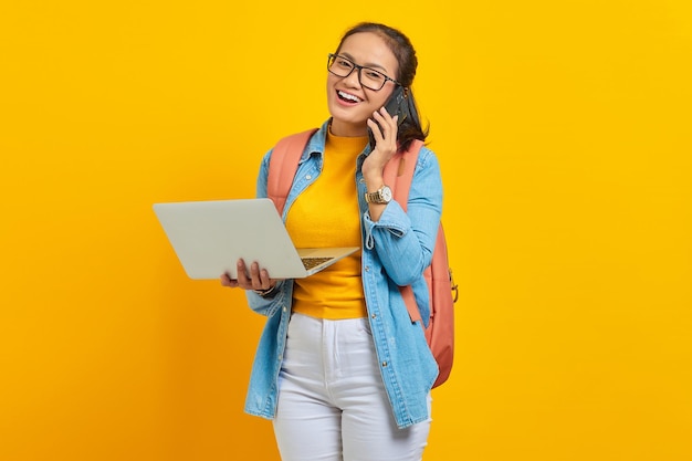 Retrato de uma jovem estudante asiática alegre em roupas casuais com mochila falando no celular e segurando laptop isolado em fundo amarelo. Educação no conceito de universidade de faculdade