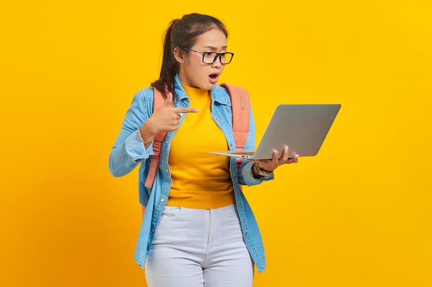 Retrato de uma jovem estudante asiática surpresa em roupas casuais com mochila apontando dedos para laptop isolado em fundo amarelo Educação no conceito de faculdade universitária