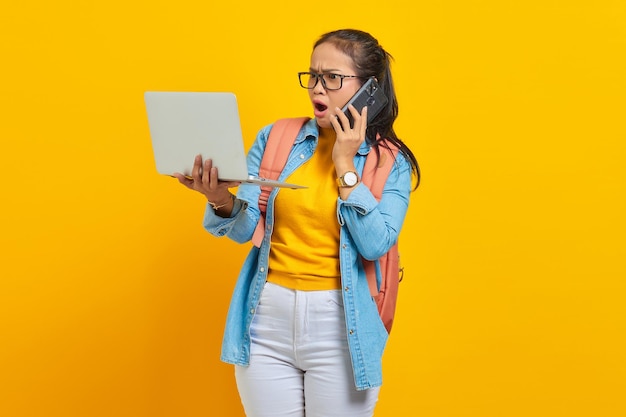 Retrato de uma jovem estudante asiática surpresa em roupas casuais com mochila falando no celular e usando laptop isolado em fundo amarelo. Educação no conceito de universidade de faculdade