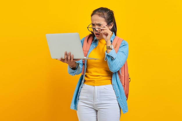 Retrato de uma jovem estudante asiática surpresa em roupas casuais com mochila usando laptop e segurando óculos com a mão isolada em fundo amarelo Educação no conceito de universidade universitária
