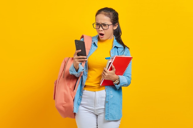 Retrato de uma jovem estudante asiática surpresa em roupas jeans com mochila usando telefone celular digitando mensagem sms e segurando livros isolados em fundo amarelo