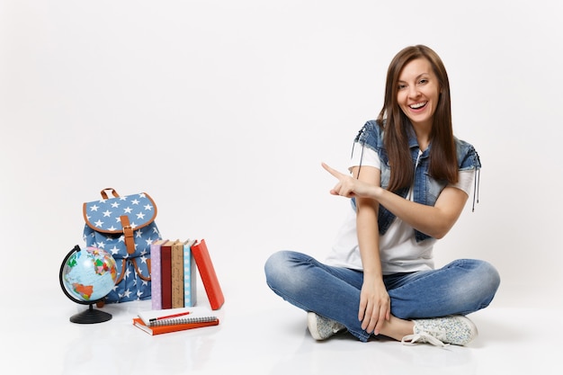 Retrato de uma jovem estudante casual rindo em roupas jeans, sentada, apontando o dedo indicador na mochila do globo, livros escolares isolados