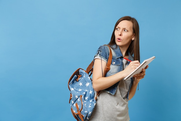 Retrato de uma jovem estudante duvidosa preocupado em t-shirt cinza, roupas jeans com mochila olhar de lado escrevendo no caderno isolado sobre fundo azul. educação na faculdade universitária do ensino médio.