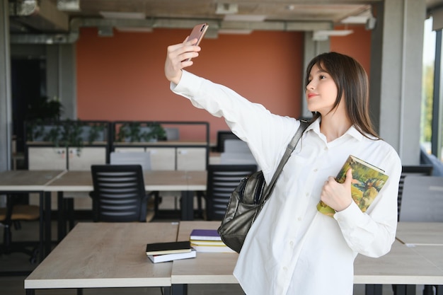 Retrato de uma jovem estudante feliz e sorridente, com cabelos longos, parada em uma sala de aula vazia, olhando para a câmera, tirando uma selfie
