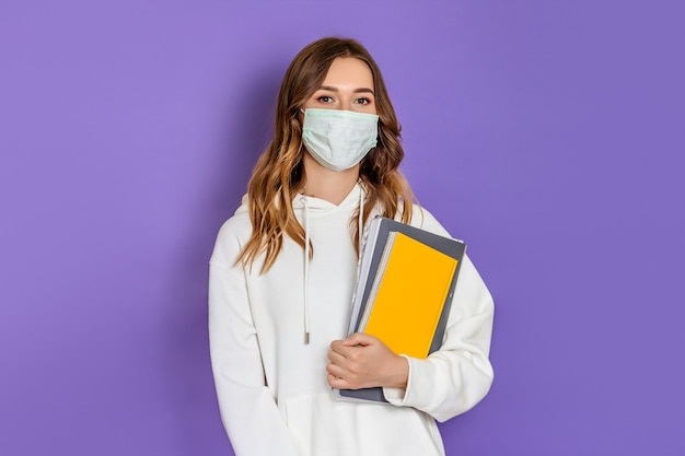 Retrato de uma jovem estudante morena com máscara cirúrgica médica usando um capuz branco em pé e olhando para a câmera isolada sobre fundo lilás