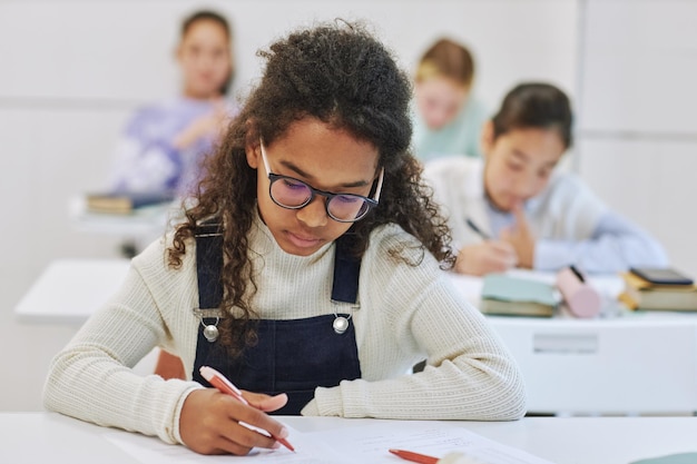 Retrato de uma jovem estudante negra sentada à mesa na sala de aula da escola e fazendo teste