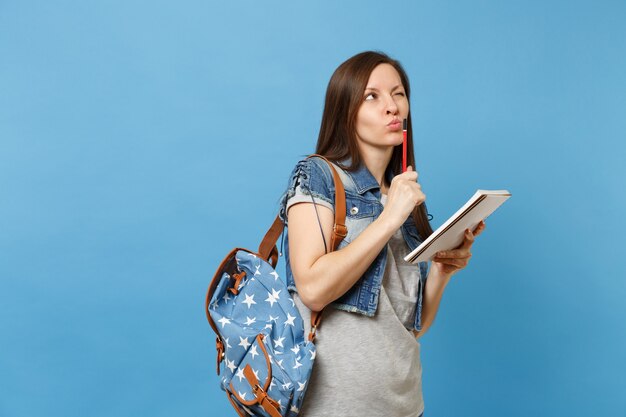 Retrato de uma jovem estudante pensativa em roupas jeans com mochila, fazendo o exame pensando em teste, segurando o lápis do caderno isolado sobre fundo azul. Educação na faculdade universitária do ensino médio.