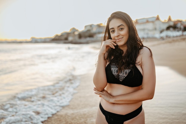 Retrato de uma jovem europeia feliz em trajes de banho pretos posando de pé na praia na costa do oceano