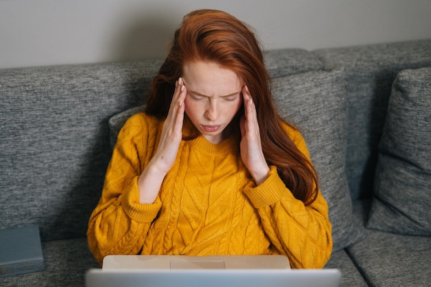 Retrato de uma jovem exausta com dor de cabeça enquanto trabalhava digitando no laptop sentado no sofá em casa