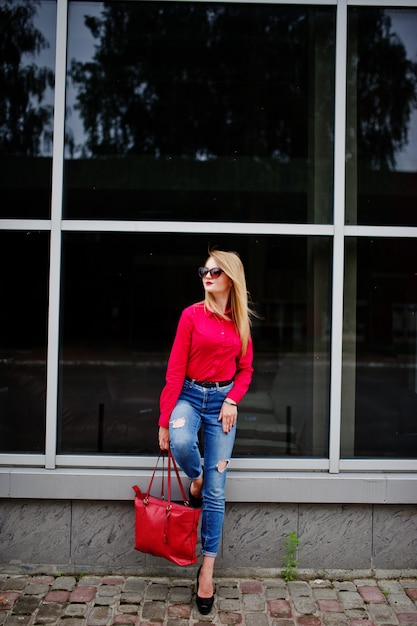 Retrato de uma jovem fabulosa na blusa vermelha e calça jeans posando com sua bolsa e óculos de sol fora do shopping