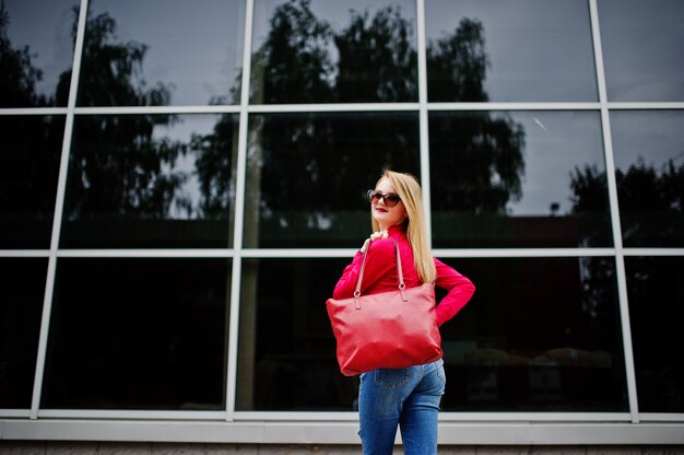 Retrato de uma jovem fabulosa na blusa vermelha e calça jeans posando com sua bolsa e óculos de sol fora do shopping