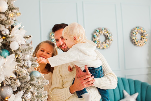 Retrato de uma jovem família perto da véspera de Natal Linda mãe com cabelos longos e encaracolados e pai atraente ajudando seu lindo filho loiro a pendurar a árvore de Natal