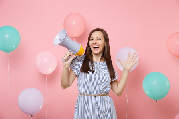 Retrato de uma jovem fascinante mulher feliz usando um vestido azul, segurando o megafone, espalhando as mãos no fundo rosa brilhante com balões de ar coloridos. Festa de feriado de aniversário, pessoas sinceras emotivas