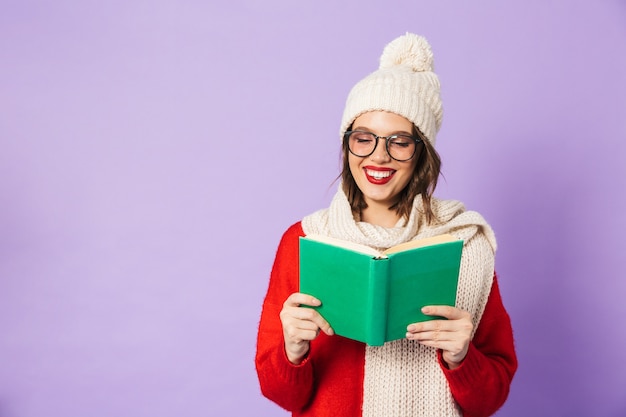 Retrato de uma jovem feliz animado com chapéu de inverno isolado sobre o livro de leitura de parede roxa.