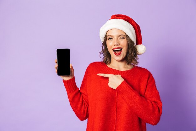 Retrato de uma jovem feliz animado usando chapéu de Natal isolado sobre a parede roxa, mostrando o visor do telefone móvel.