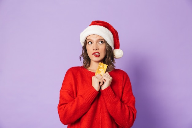 Retrato de uma jovem feliz animado usando chapéu de Natal isolado sobre a parede roxa, segurando o lábio de mordida de cartão de débito.